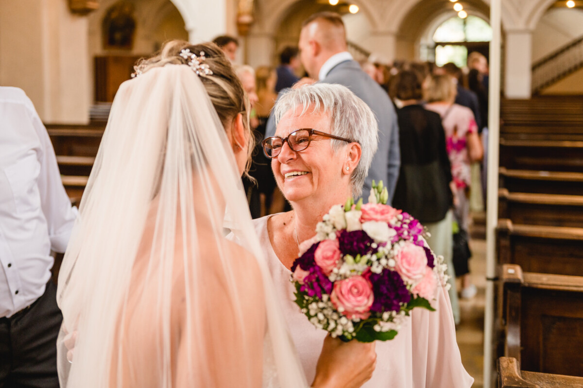 christian-lorenz.net Eine Braut mit Schleier hält einen bunten Blumenstrauß in der Hand und lächelt eine ältere Frau mit Brille in einer Kirche an.