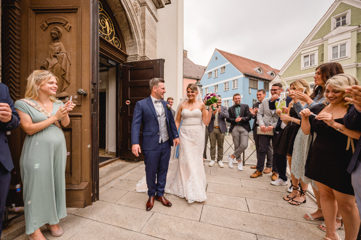 christian-lorenz.net Ein Brautpaar verlässt Händchen haltend eine Kirche. Sie sind umgeben von lächelnden Gästen in festlicher Kleidung, von denen einige klatschen und andere Getränke in der Hand halten, auf einem gepflasterten Gehweg.
