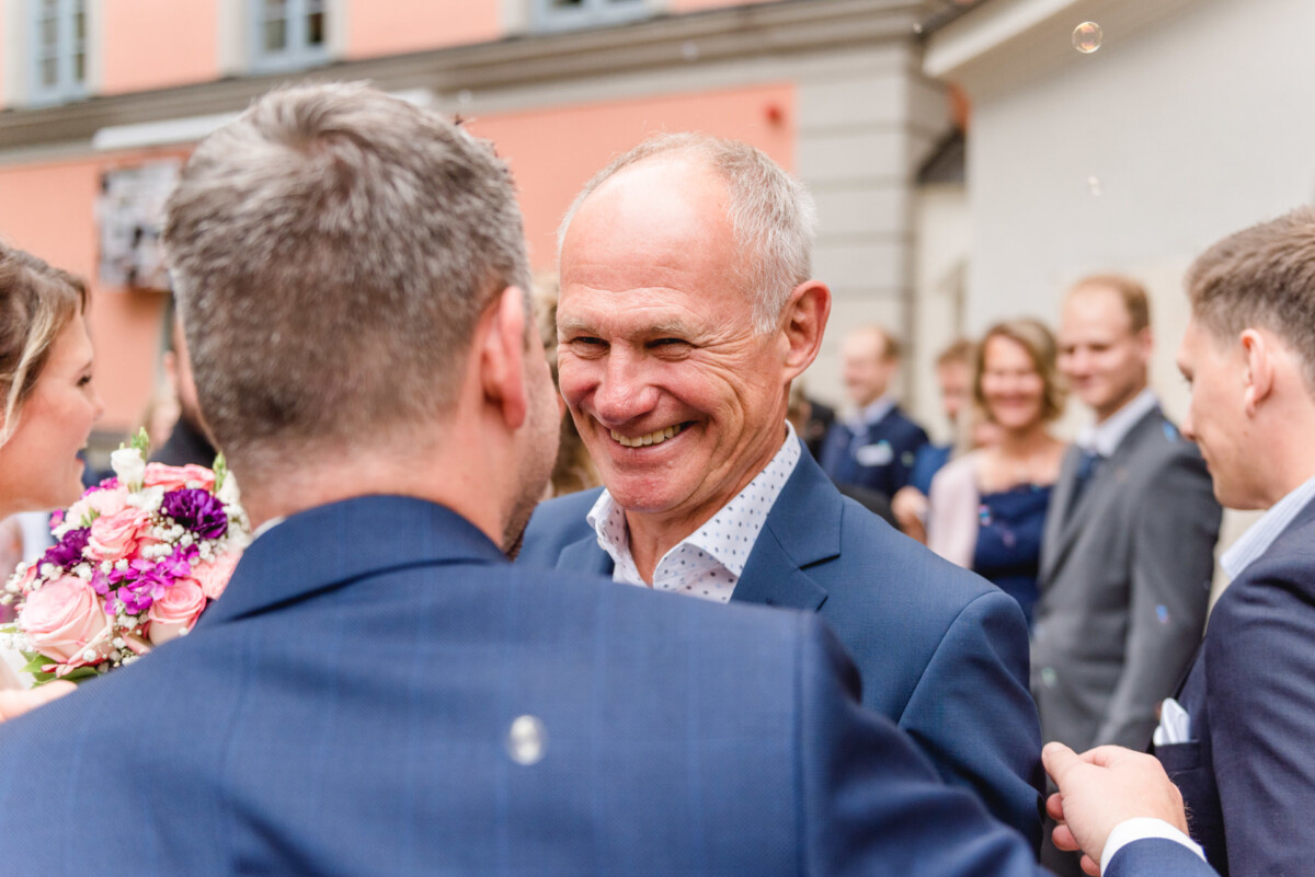 christian-lorenz.net Ein lächelnder Mann im Anzug, umgeben von Menschen in Abendgarderobe, steht im Freien in der Nähe eines Gebäudes mit einer rosa Wand.