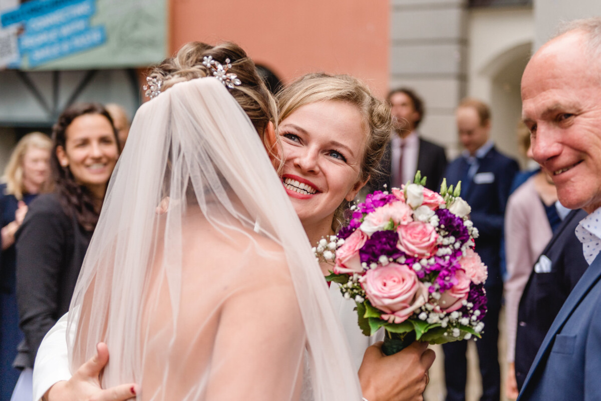 christian-lorenz.net Die Braut umarmt eine lächelnde Frau, die einen Strauß rosa und lila Blumen hält, umgeben von Gästen bei einer Hochzeitsfeier im Freien.