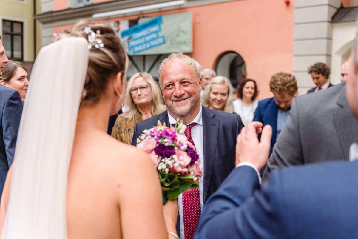 christian-lorenz.net Eine Braut mit einem Blumenstrauß steht einem lächelnden älteren Mann im Anzug gegenüber. Im Hintergrund steht eine Gruppe von Menschen vor einem Gebäude mit orangefarbenen und beigen Wänden.