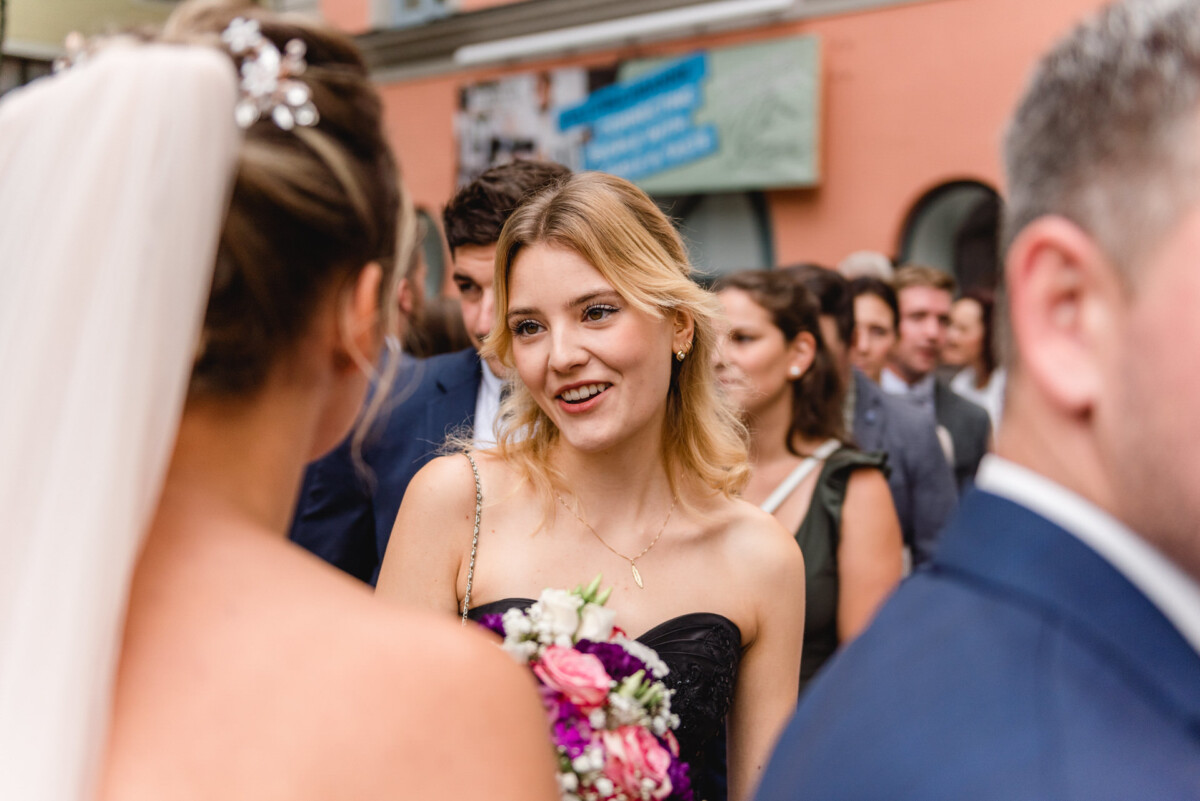 christian-lorenz.net Eine Frau in Abendkleid lächelt und hält einen Blumenstrauß in der Hand, umgeben von Menschen bei einer Veranstaltung, die wie eine gesellschaftliche Veranstaltung oder Zusammenkunft aussieht.