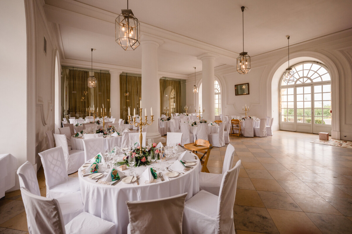 christian-lorenz.net Eleganter Speisesaal für eine Veranstaltung mit runden Tischen mit weißen Tischdecken, dekorativen Tischdekorationen und Stühlen. Große Fenster und Kronleuchter unterstreichen die helle Atmosphäre.