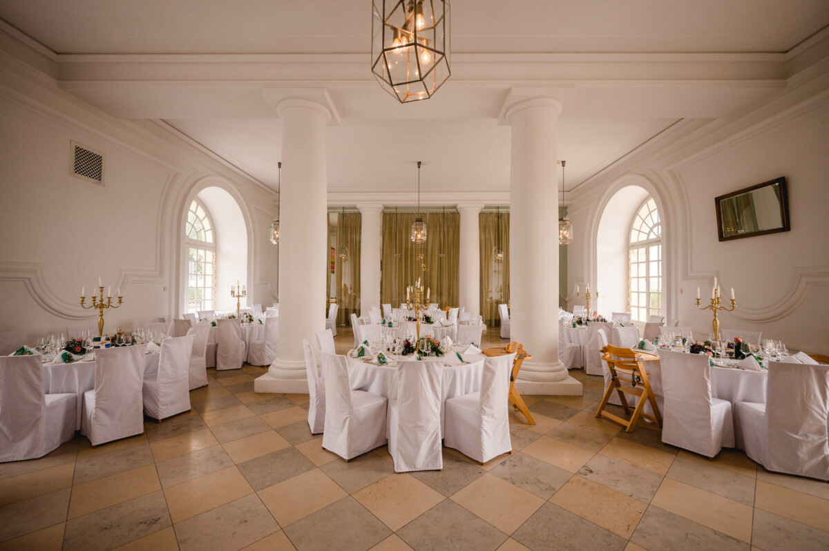 christian-lorenz.net Ein Bankettsaal mit runden Tischen mit weißen Tischdecken und Stühlen mit weißem Stoffbezug. Der Raum besticht durch große Säulen, Bogenfenster und dekorative Kronleuchter.