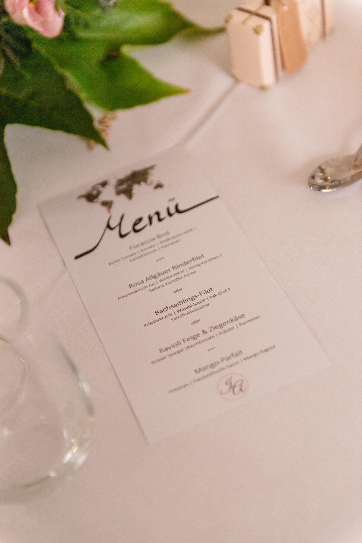 christian-lorenz.net Eine Speisekarte auf einer weißen Tischdecke neben einem Glas und einem Löffel. Darauf stehen Gerichte wie „Rosa Allgäuer Rinderfilet“ und „Ravioli Feine & Ziegenkäse“. Dekoratives Laub und rosa Blüten sind zu sehen.