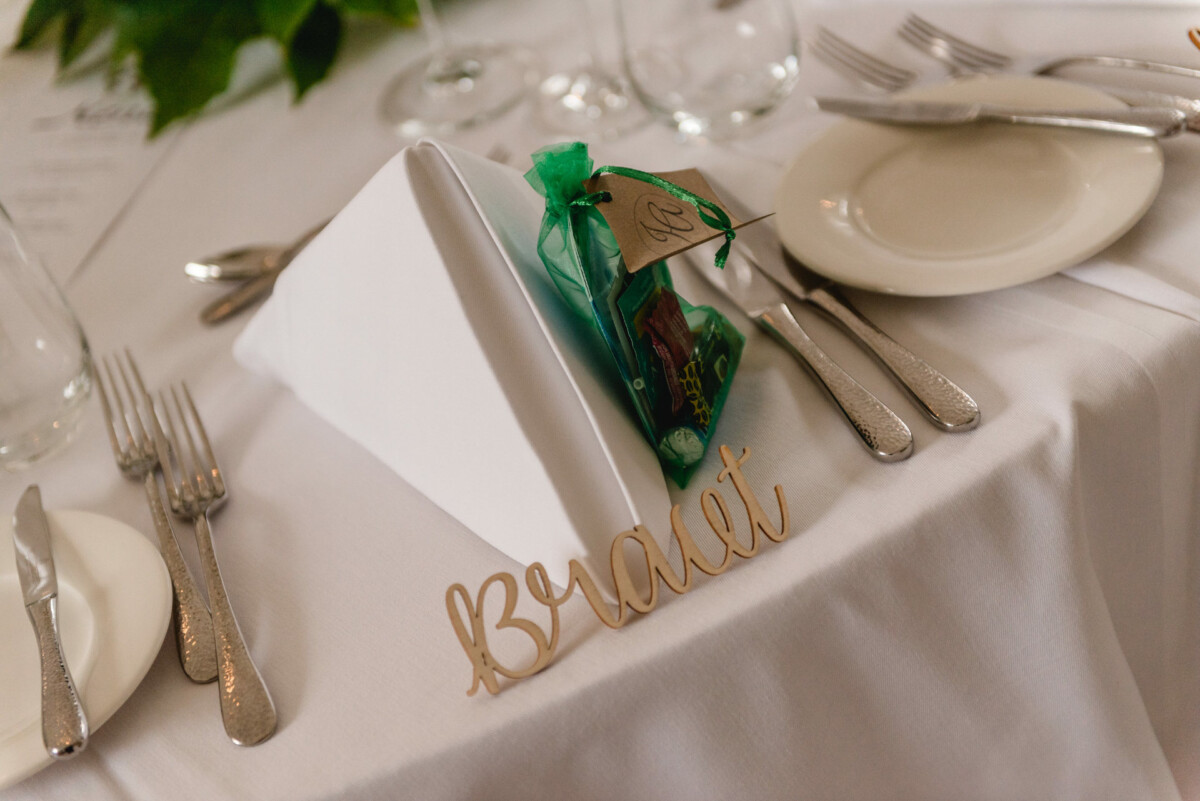christian-lorenz.net Eine weiße Tischdecke mit gefalteter Serviette, Besteck und einer kleinen grünen Geschenktüte. Am Rand befindet sich ein Namensschild mit der Aufschrift „Brad“.
