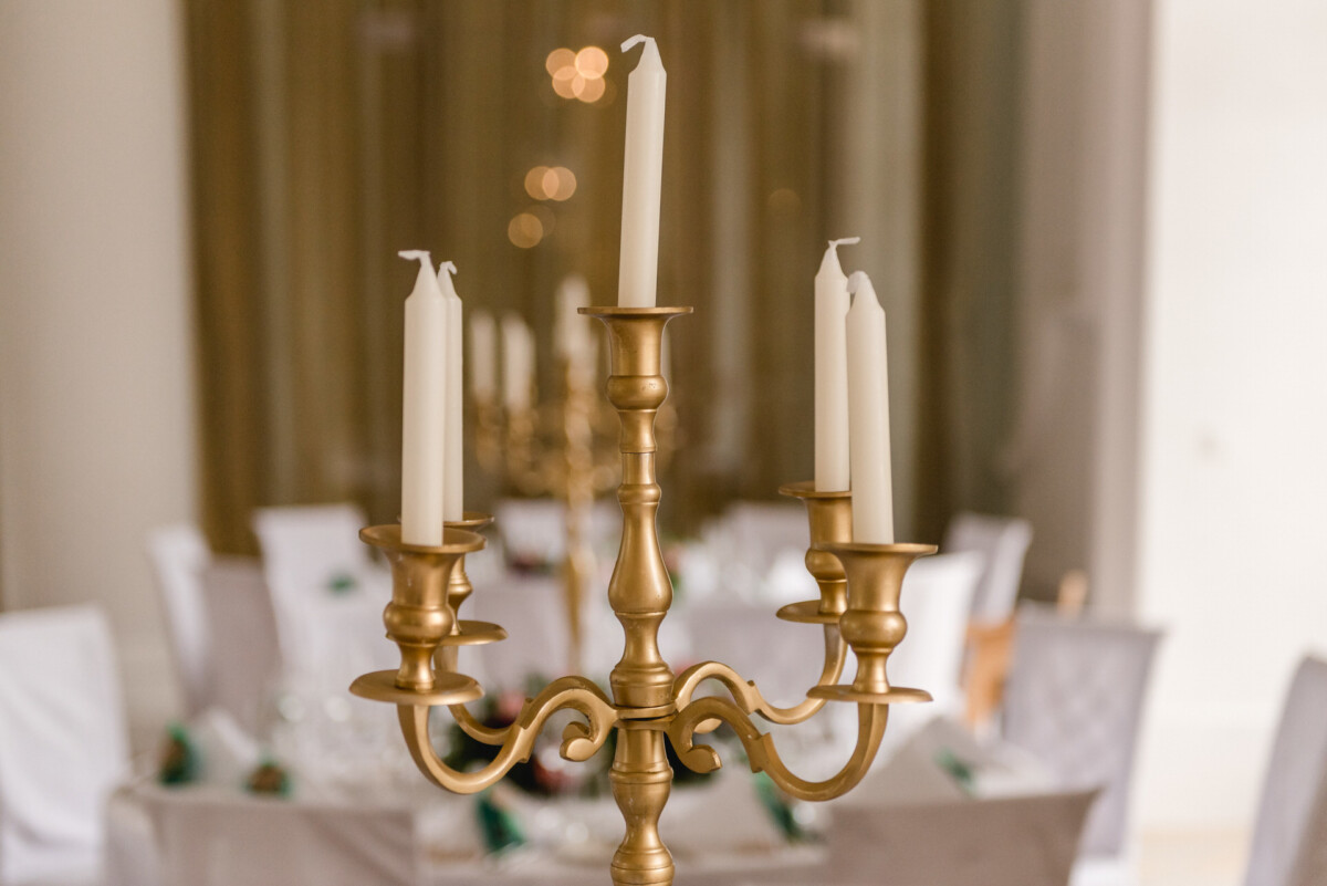 christian-lorenz.net In der Mitte steht ein goldener Kandelaber mit fünf unbeleuchteten weißen Kerzen. Der unscharfe Hintergrund zeigt elegant gedeckte Tische mit weißen Tischdecken und Stuhlhussen.