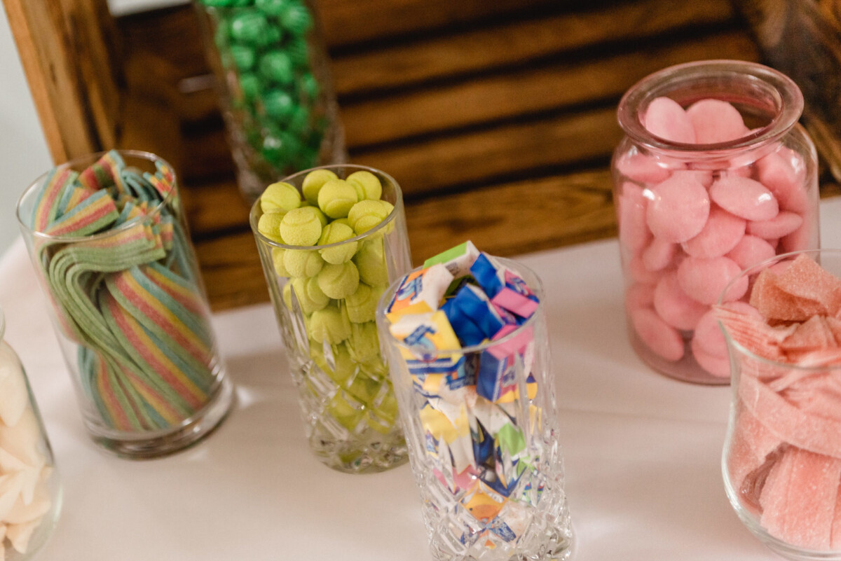 christian-lorenz.net Verschiedene Süßigkeiten in Glasbehältern, darunter gestreifte Gürtel, grüne Kugeln, eingewickelte Bonbons und rosa Bonbons, auf einem Holzständer mit einer weißen Tischdecke.