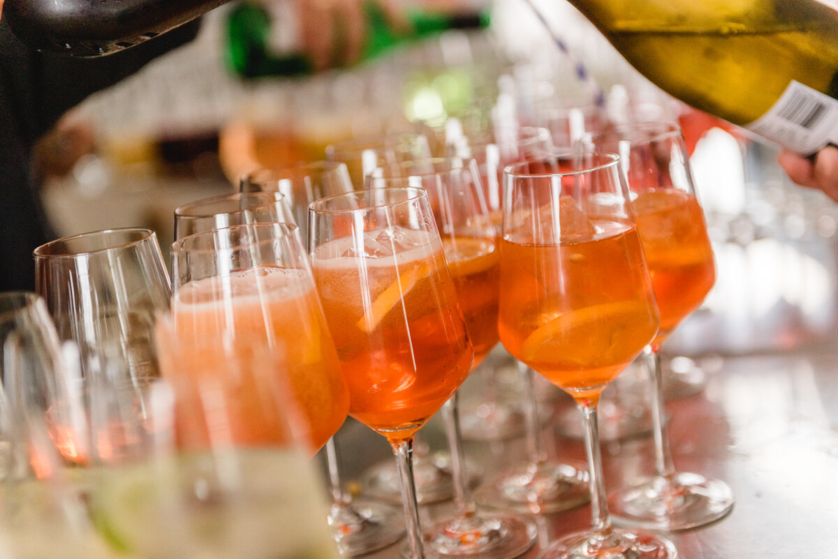 christian-lorenz.net Mehrere Gläser Orangencocktails mit Eis und Orangenscheiben stehen auf einer Theke, eine Hand schenkt aus einer Flasche nach.