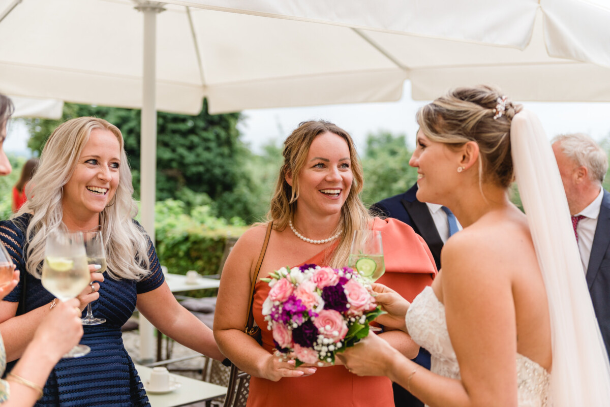 christian-lorenz.net Eine Braut im Brautkleid hält einen Blumenstrauß in der Hand und unterhält sich mit drei Frauen unter großen Sonnenschirmen. Zwei Frauen halten Weingläser, während alle lächeln. Im Hintergrund ist Grün zu sehen.