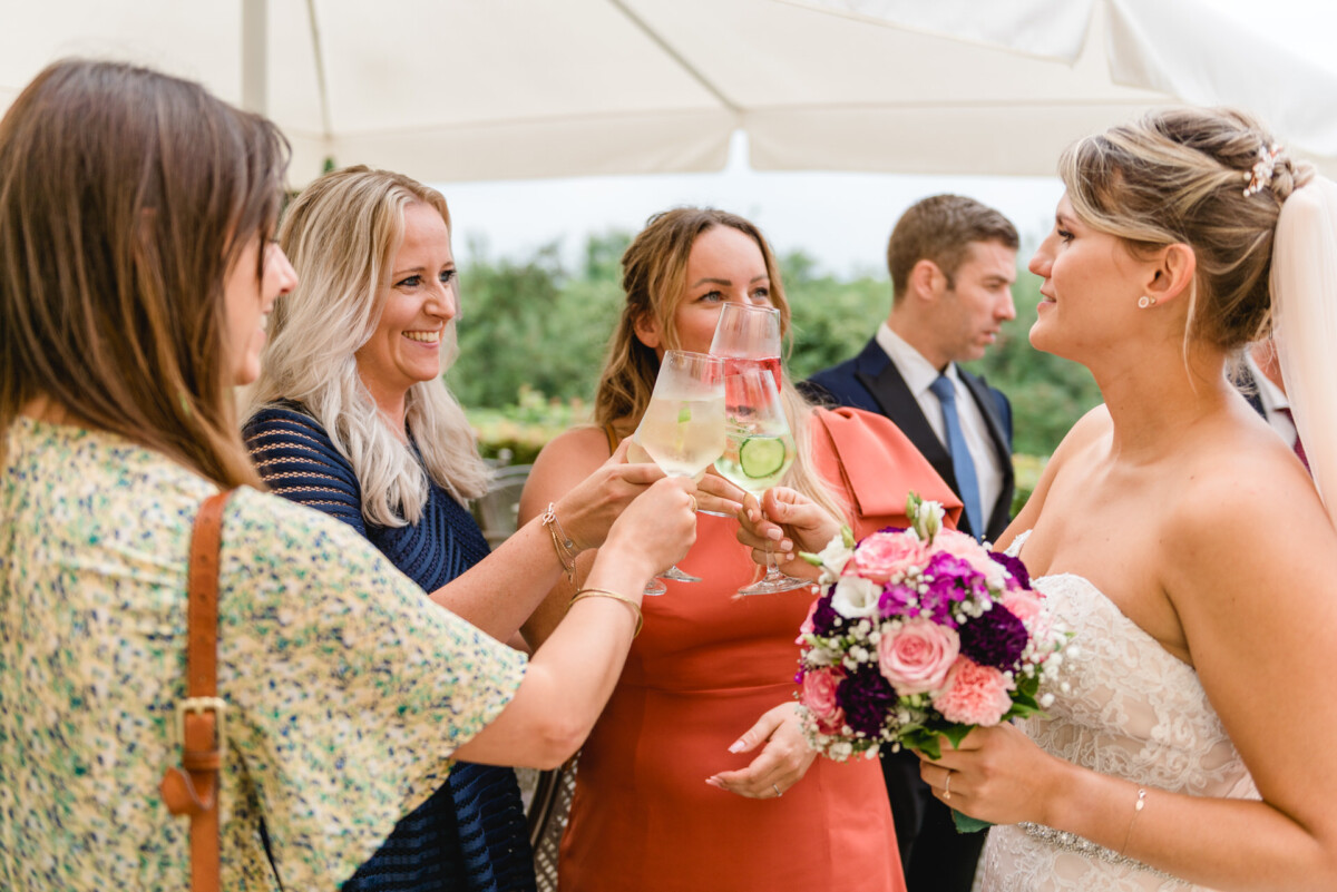 christian-lorenz.net Eine Braut hält einen Blumenstrauß in der Hand und stößt mit drei Gästen bei einer Veranstaltung im Freien an. Sie lächeln unter einem großen Sonnenschirm.