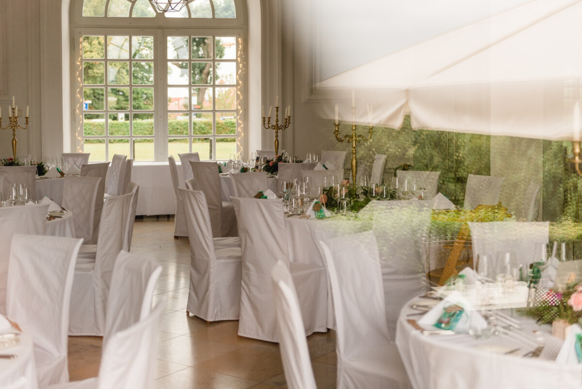 christian-lorenz.net Elegant eingerichtetes Esszimmer mit runden, weiß gedeckten Tischen, Stühlen mit Hussen, Kandelabern und großen Fenstern, durch die man einen schönen Blick auf das Grün draußen hat.