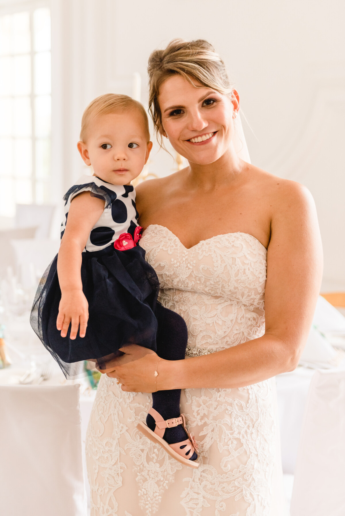 christian-lorenz.net Eine Frau in einem weißen Spitzenkleid hält ein Kleinkind in einem gepunkteten Kleid mit einer roten Schleife. Sie stehen lächelnd im Haus.