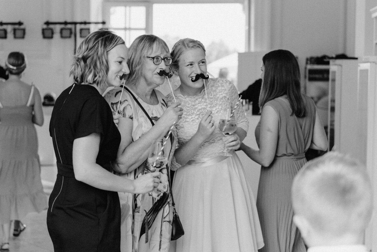 christian-lorenz.net Drei Personen posieren mit falschen Schnurrbärten und Getränken bei einer gesellschaftlichen Veranstaltung. Andere sind im Hintergrund zu sehen. Schwarz-Weiß-Foto.