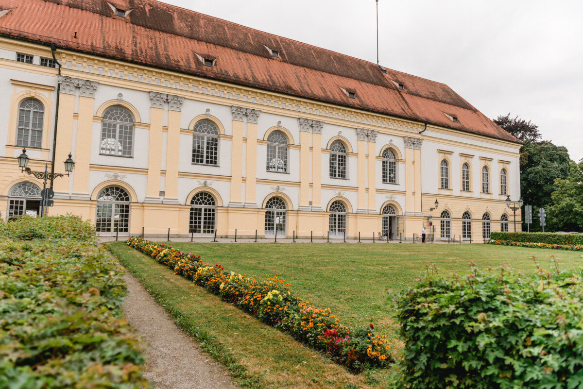 christian-lorenz.net Ein großes, gelb-weißes Gebäude mit rotem Dach und Bogenfenstern. Es ist von einem gepflegten Garten mit einem Weg und blühenden Pflanzen umgeben.