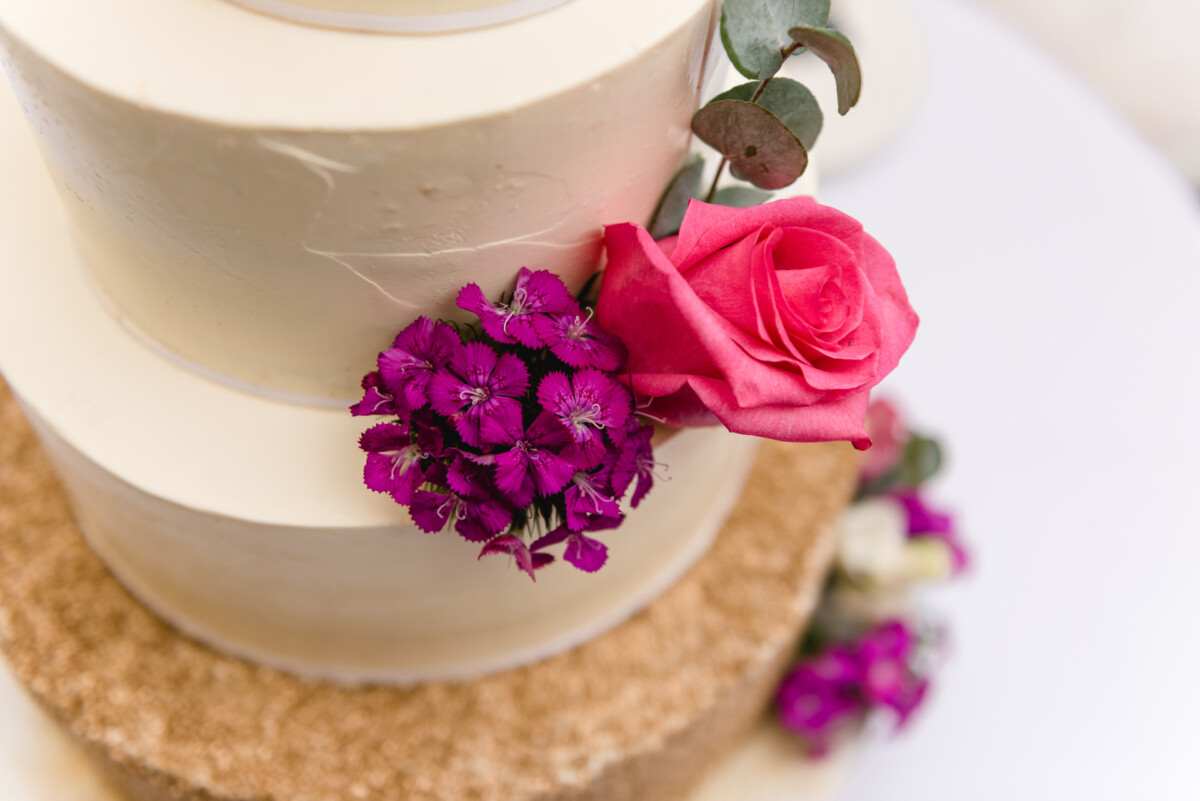 christian-lorenz.net Eine mehrstöckige Torte mit weißem Zuckerguss, verziert mit einer rosa Rose und lila Blumen, und einem mit braunen Streuseln bedeckten Boden.