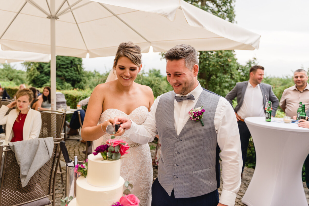 christian-lorenz.net Ein lächelndes Brautpaar schneidet bei einem Empfang im Freien eine weiße, mit rosa Blumen geschmückte Torte an. Gäste sitzen und stehen in der Nähe unter Sonnenschirmen.