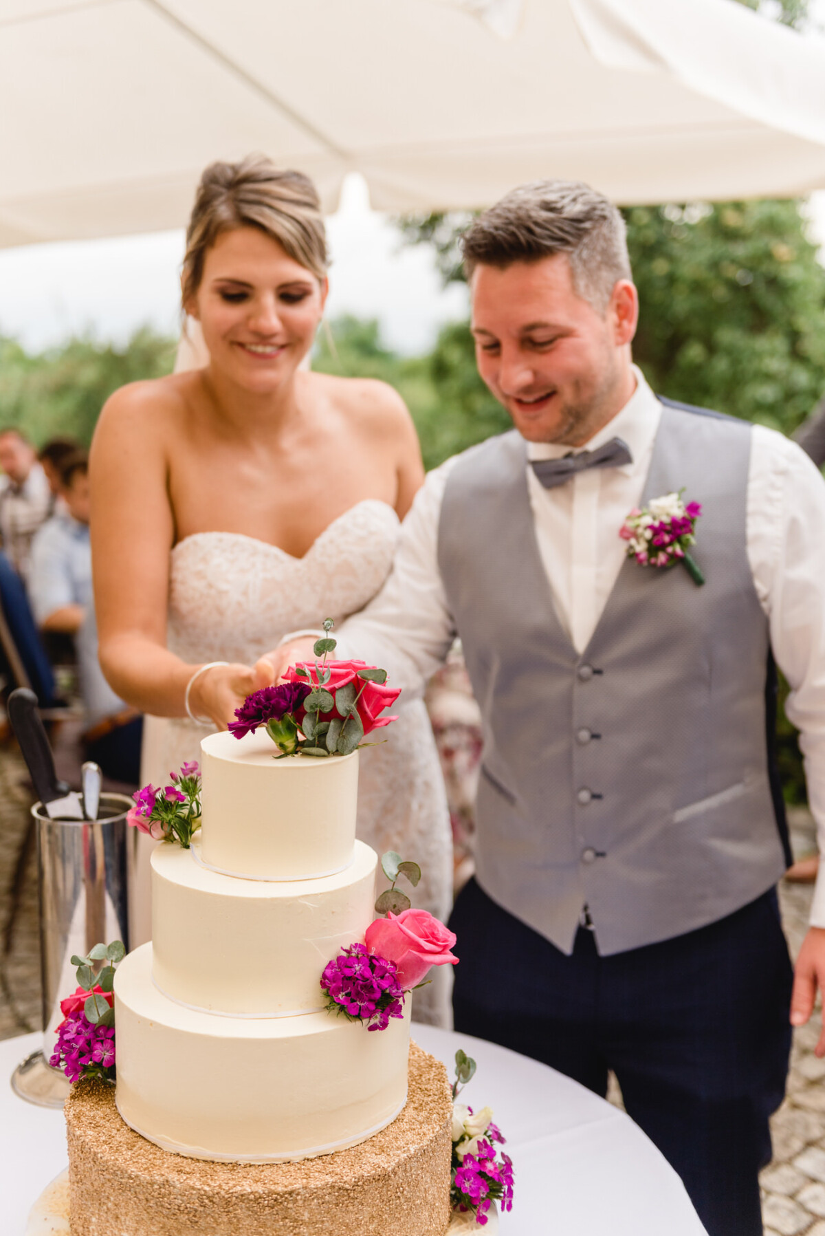 christian-lorenz.net Bei einer Veranstaltung im Freien schnitten Braut und Bräutigam eine dreistöckige Hochzeitstorte an, die mit rosa Blumen und Grünpflanzen dekoriert war.