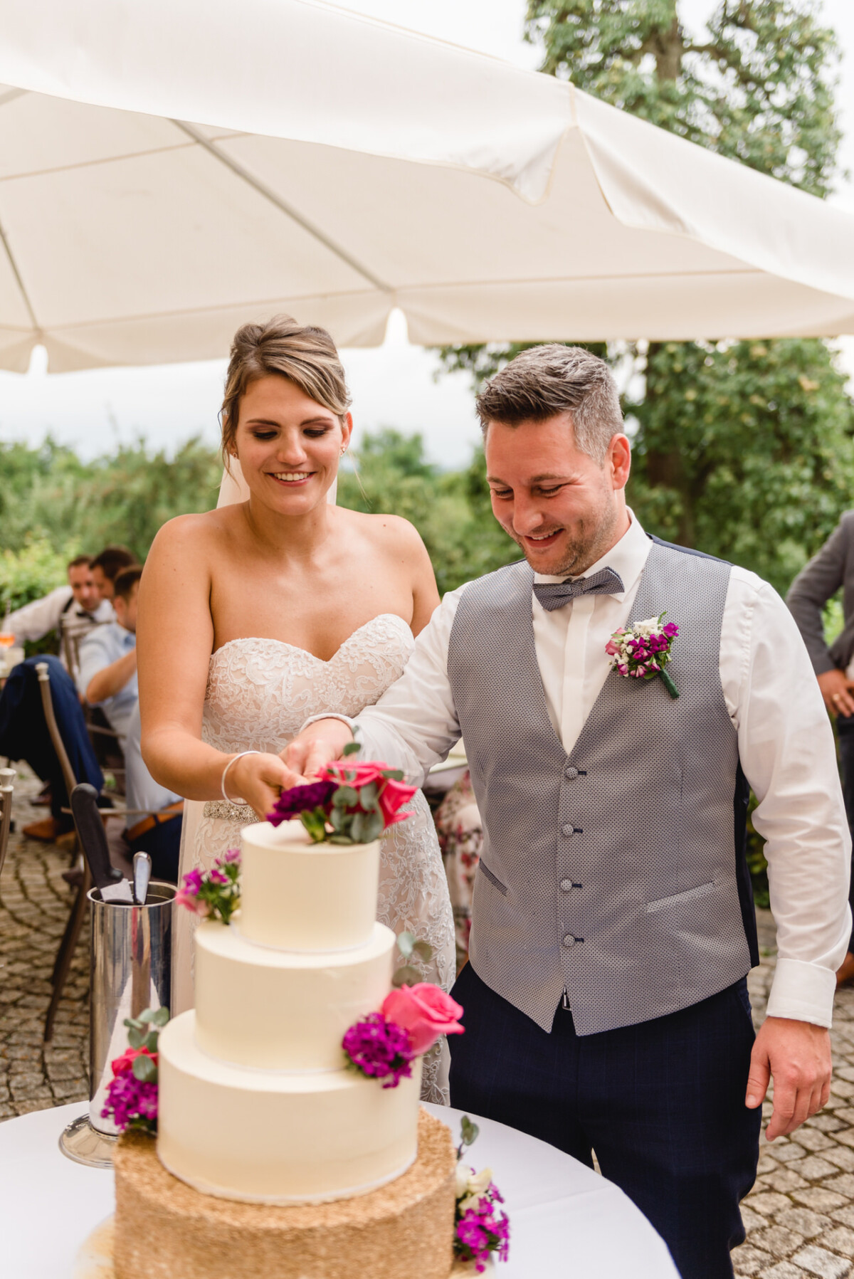 christian-lorenz.net Braut und Bräutigam schneiden im Freien eine mehrstöckige Hochzeitstorte an. Die Braut trägt ein weißes Kleid, der Bräutigam Weste und Fliege. Die Torte ist mit rosa und lila Blumen dekoriert.