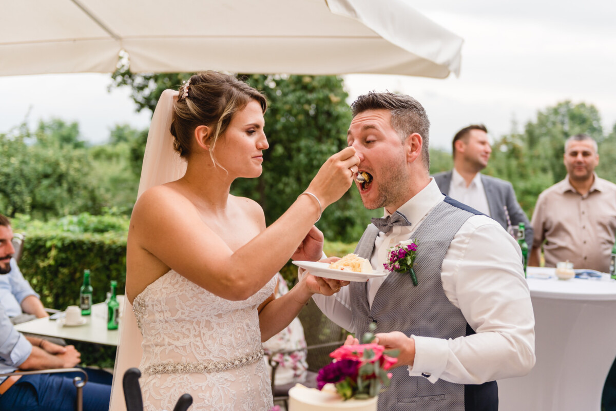 christian-lorenz.net Bei einer Hochzeitsfeier im Freien füttert die Braut den Bräutigam spielerisch mit der Torte. Beide lächeln. Im Hintergrund Menschen und ein Tisch mit Blumen.