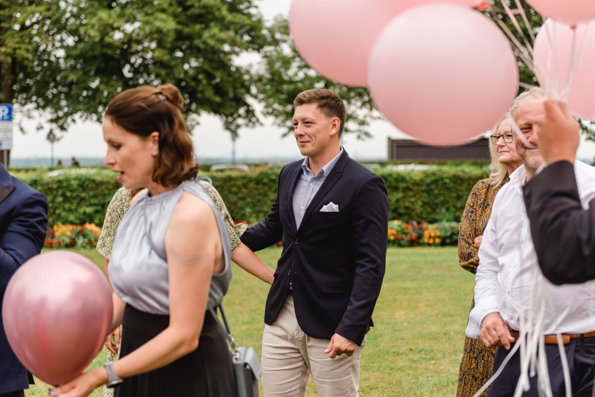 christian-lorenz.net Eine Gruppe von Menschen steht im Freien, einige halten rosa Luftballons. Im Vordergrund stehen ein Mann in einem dunklen Blazer und eine Frau in einem silbernen Top. Im Hintergrund sind Bäume und Büsche zu sehen.