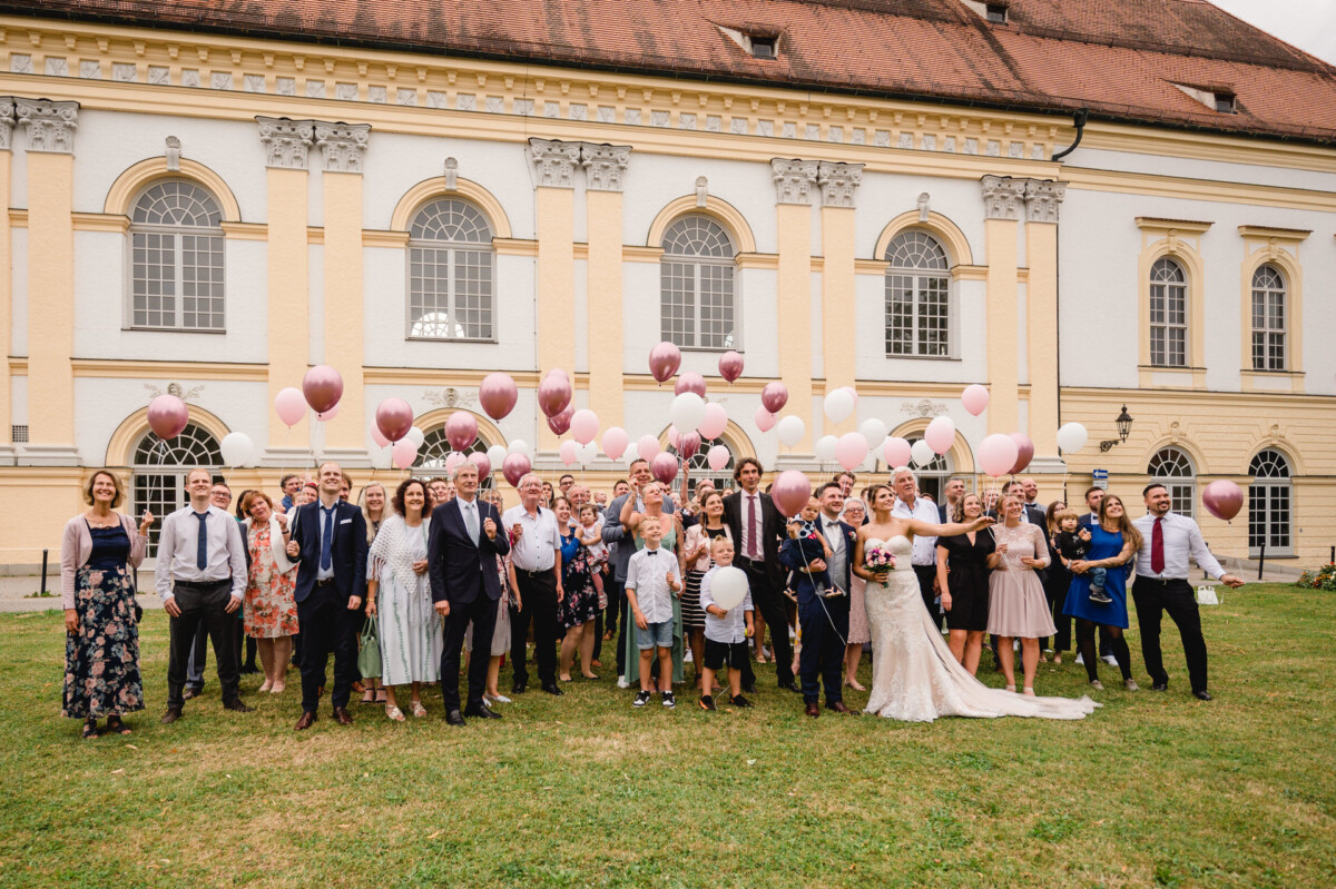 christian-lorenz.net Eine Hochzeitsgesellschaft steht auf einer Wiese vor einem großen Gebäude mit Bogenfenstern. Die Gäste halten rosa und weiße Luftballons in den Händen, von denen einige in die Luft steigen.