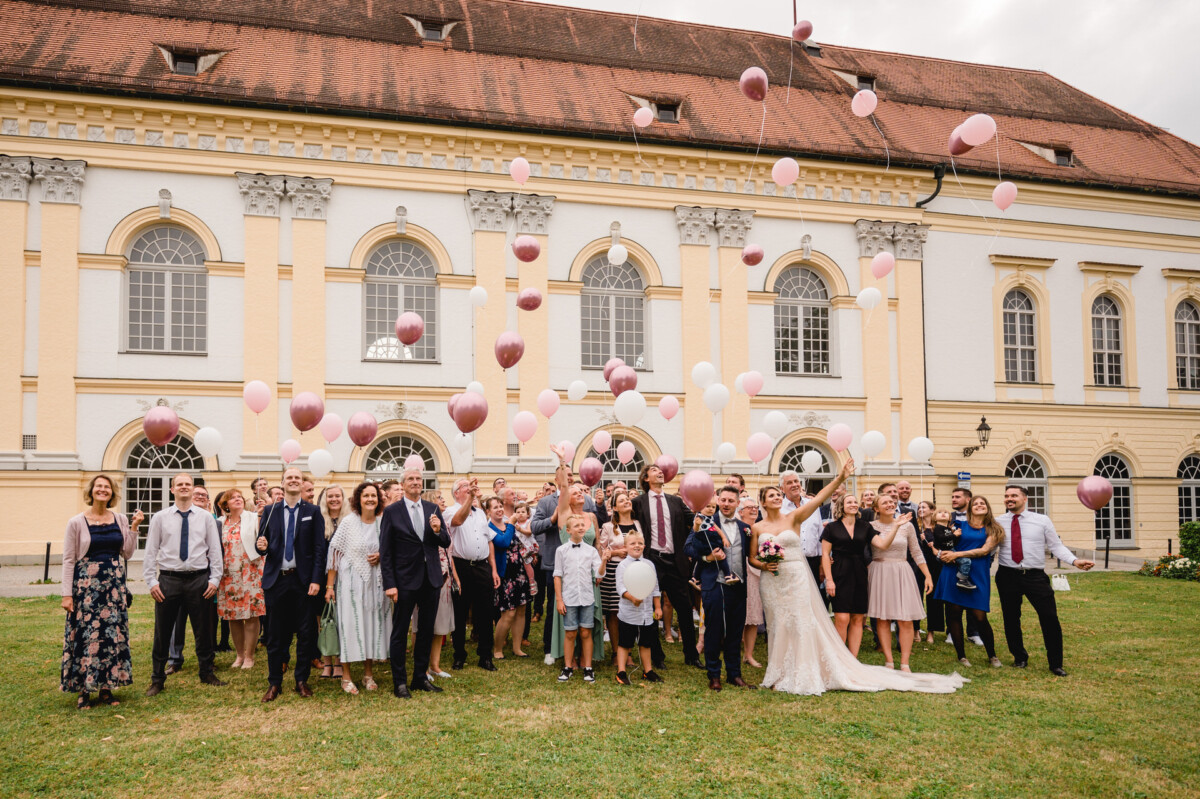 christian-lorenz.net Eine Gruppe von Hochzeitsgästen lässt vor einem großen historischen Gebäude rosa und weiße Luftballons steigen.