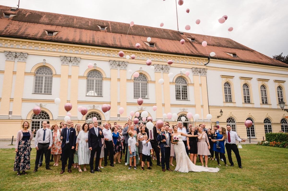 christian-lorenz.net Eine große Gruppe von Menschen, darunter ein Brautpaar, feiert vor einem historischen Gebäude. Sie lassen rosa und weiße Luftballons in den Himmel steigen.