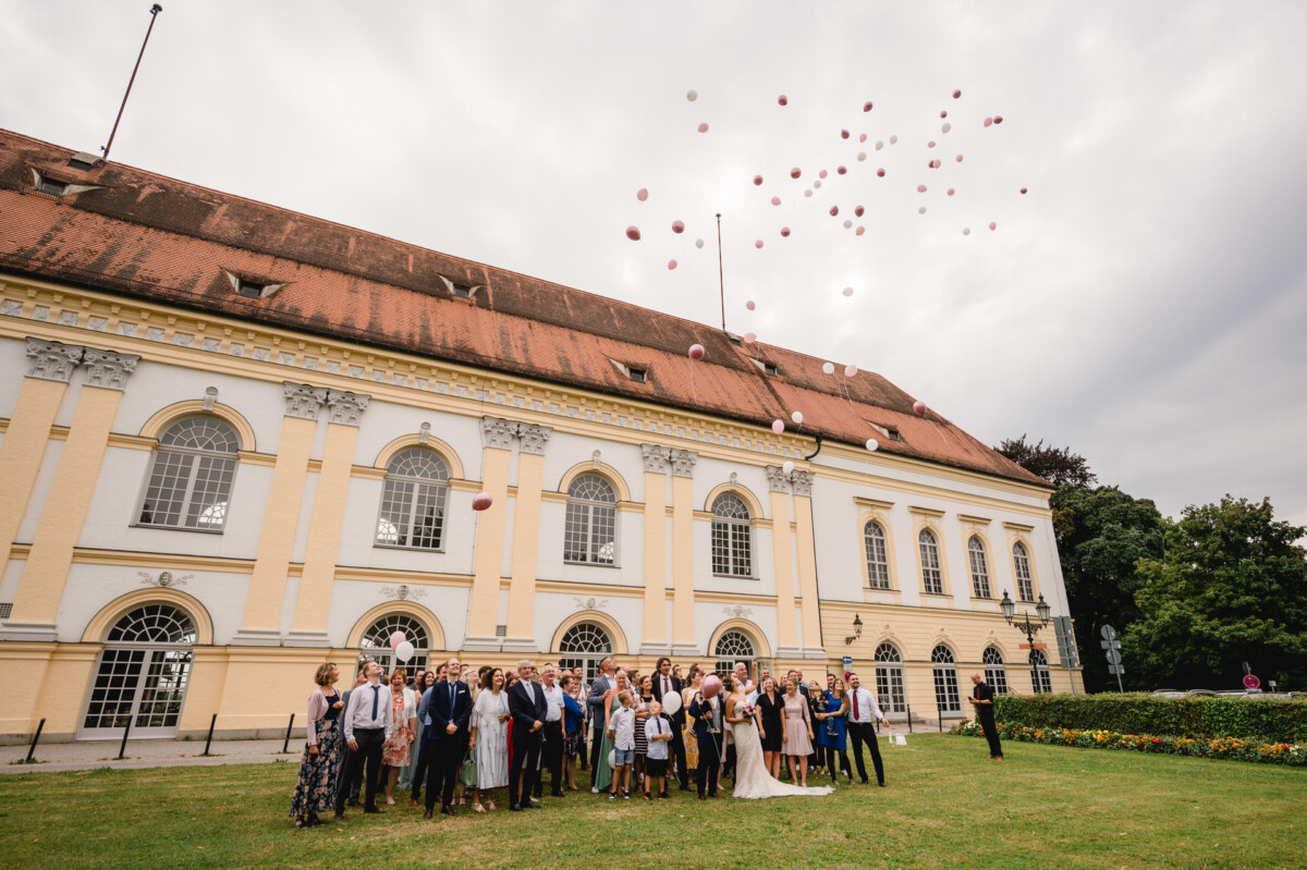 christian-lorenz.net Eine große Gruppe von Menschen steht auf einer Wiese vor einem historischen Gebäude und lässt rosa und weiße Luftballons in den bewölkten Himmel steigen.