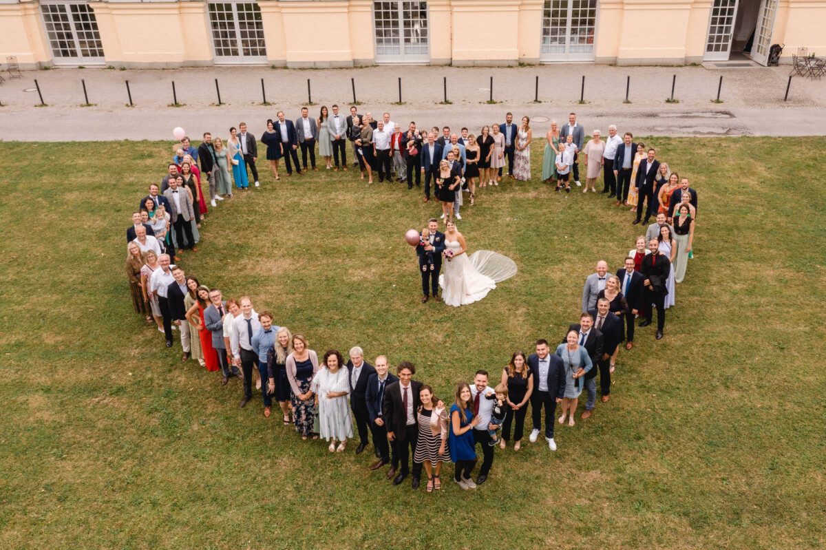 Eine große Gruppe von Menschen hat sich herzförmig auf einer Wiese angeordnet und umringt ein Paar in Hochzeitskleidung, das rosa Luftballons hält. Im Hintergrund ist ein großes Gebäude zu sehen.