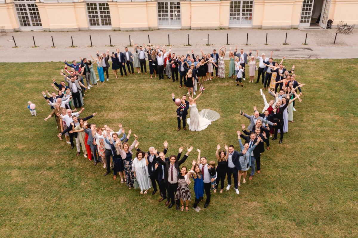 Luftaufnahme einer Hochzeitsgruppe, die in Herzform auf einem Rasen posiert, mit dem Paar in der Mitte, vor einem großen Gebäude.