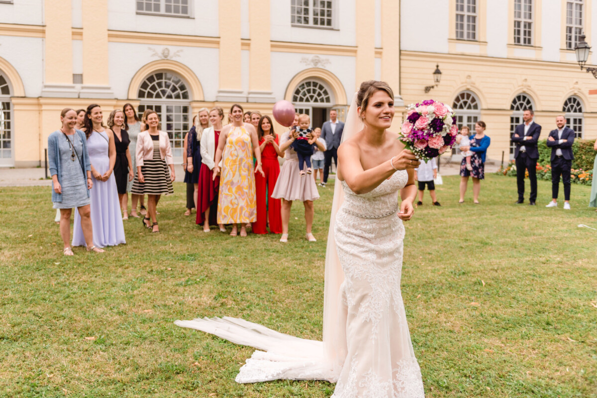 christian-lorenz.net Eine Braut in einem weißen Kleid hält einen Blumenstrauß in der Hand und bereitet sich darauf vor, ihn in die Luft zu werfen. Hinter ihr steht eine Gruppe von Menschen auf einer Rasenfläche vor einem großen Gebäude mit Bogenfenstern.