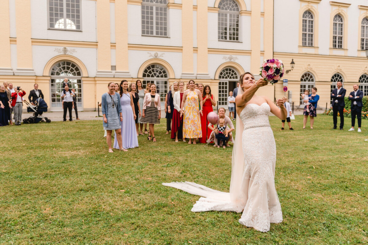 christian-lorenz.net Eine Braut in einem weißen Kleid bereitet sich darauf vor, ihren Blumenstrauß einer Gruppe wartender Gäste vor einem großen Gebäude mit Bogenfenstern zuzuwerfen.