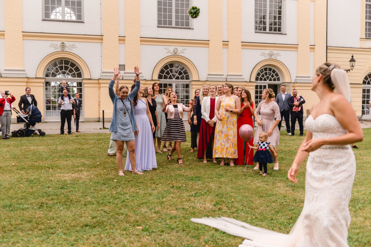 christian-lorenz.net Eine Braut in einem weißen Kleid bereitet sich darauf vor, ihren Brautstrauß einer Gruppe aufgeregter Frauen zuzuwerfen, die sich auf einer Wiese vor einem großen Gebäude versammelt haben.