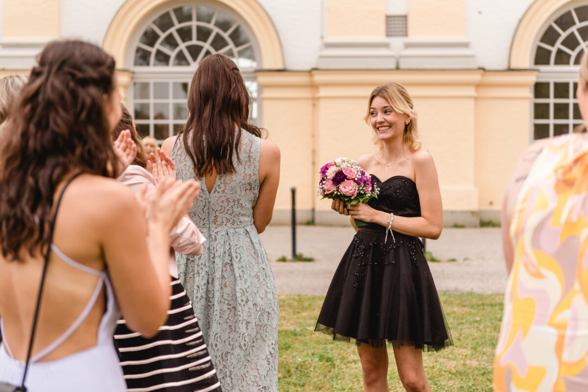 christian-lorenz.net Eine Frau im schwarzen Kleid hält einen Blumenstrauß in der Hand und lächelt, während sie draußen vor einem gelben Gebäude inmitten einer Gruppe applaudierender Menschen steht.