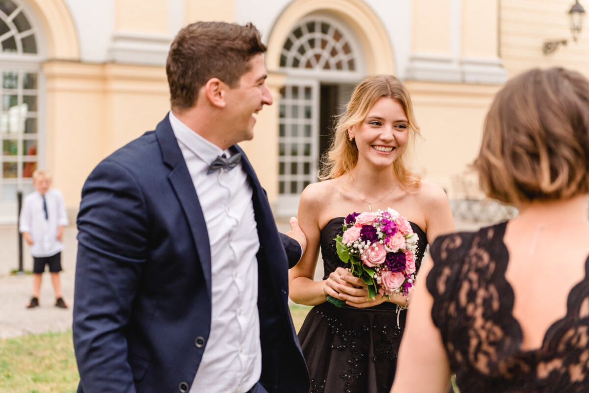 christian-lorenz.net Eine lächelnde Frau in einem schwarzen Kleid hält einen Blumenstrauß in der Hand und steht mit zwei Personen im Freien. Im Hintergrund ist in der Nähe eines Gebäudes mit Bogenfenstern ein Kind zu sehen.