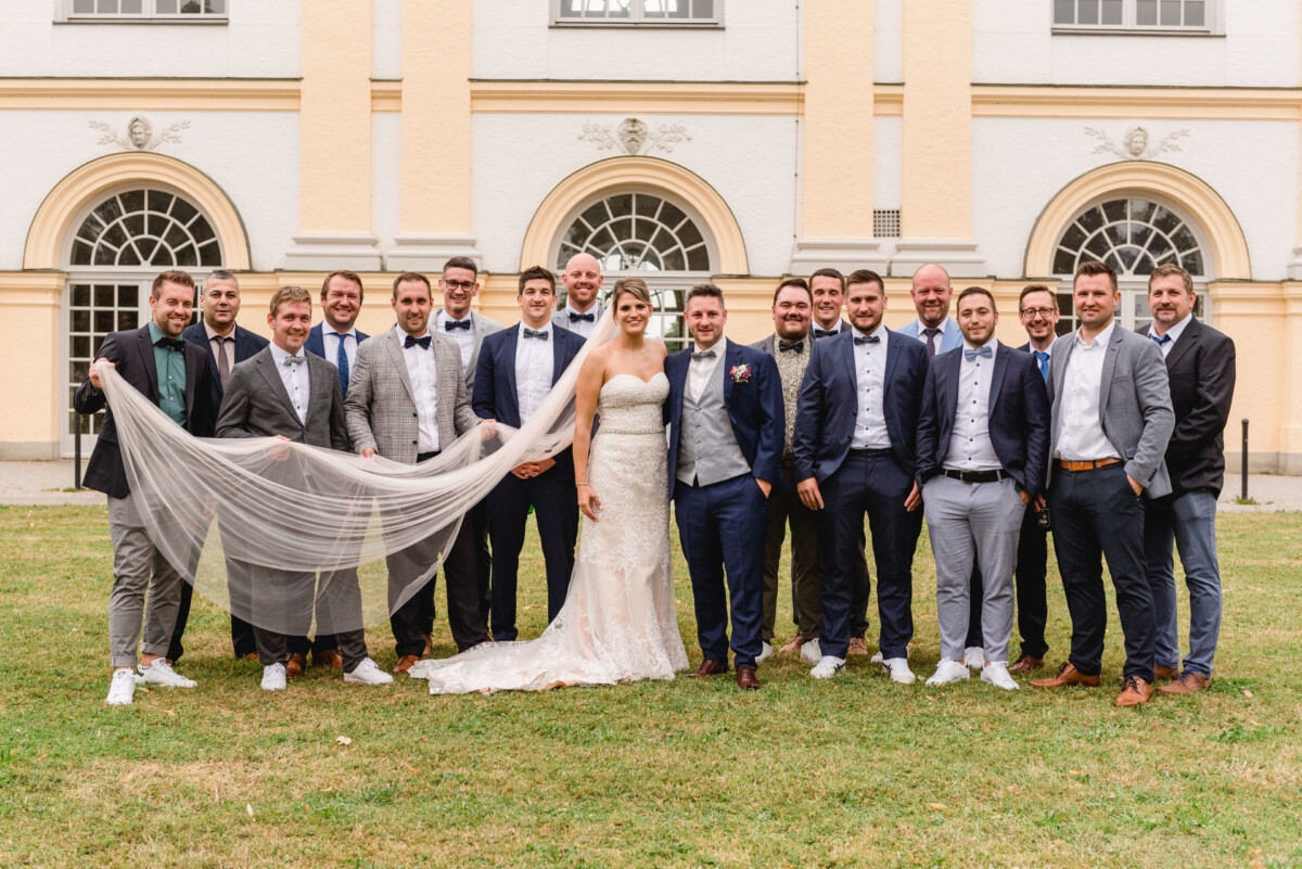 christian-lorenz.net Ein Brautpaar steht mit einer Gruppe von Männern in Anzügen und Turnschuhen vor einem Gebäude. Ein Mann hält den Schleier der Braut.