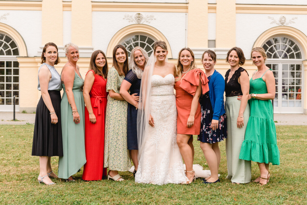 christian-lorenz.net Eine Braut in einem weißen Kleid steht mit zehn Frauen in bunten Kleidern im Freien und posiert für ein Gruppenfoto vor einem hellgelben Gebäude mit Bogenfenstern.