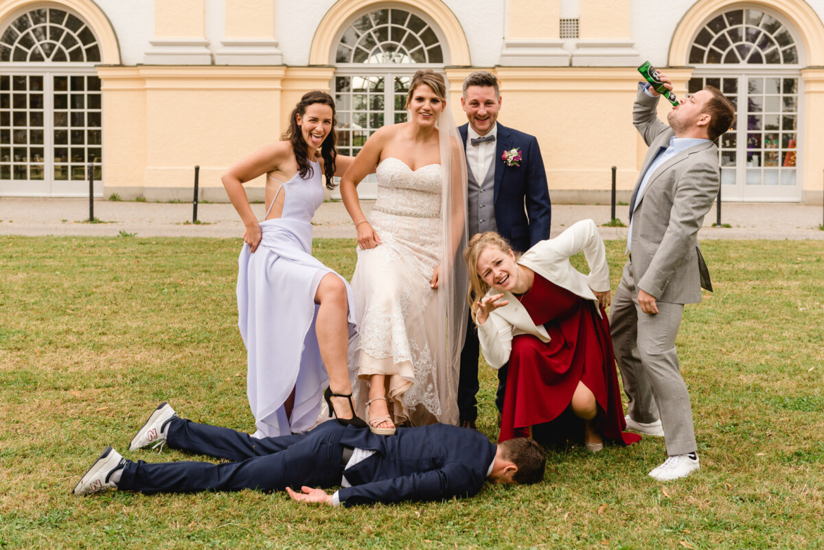christian-lorenz.net Ein Hochzeitsfoto mit Braut, Bräutigam und vier Gästen in verspielten Posen auf einer Wiese. Ein Gast liegt, andere posieren jubelnd, einer trinkt aus einer Flasche. Im Hintergrund ist ein Gebäude mit Bogenfenstern zu sehen.