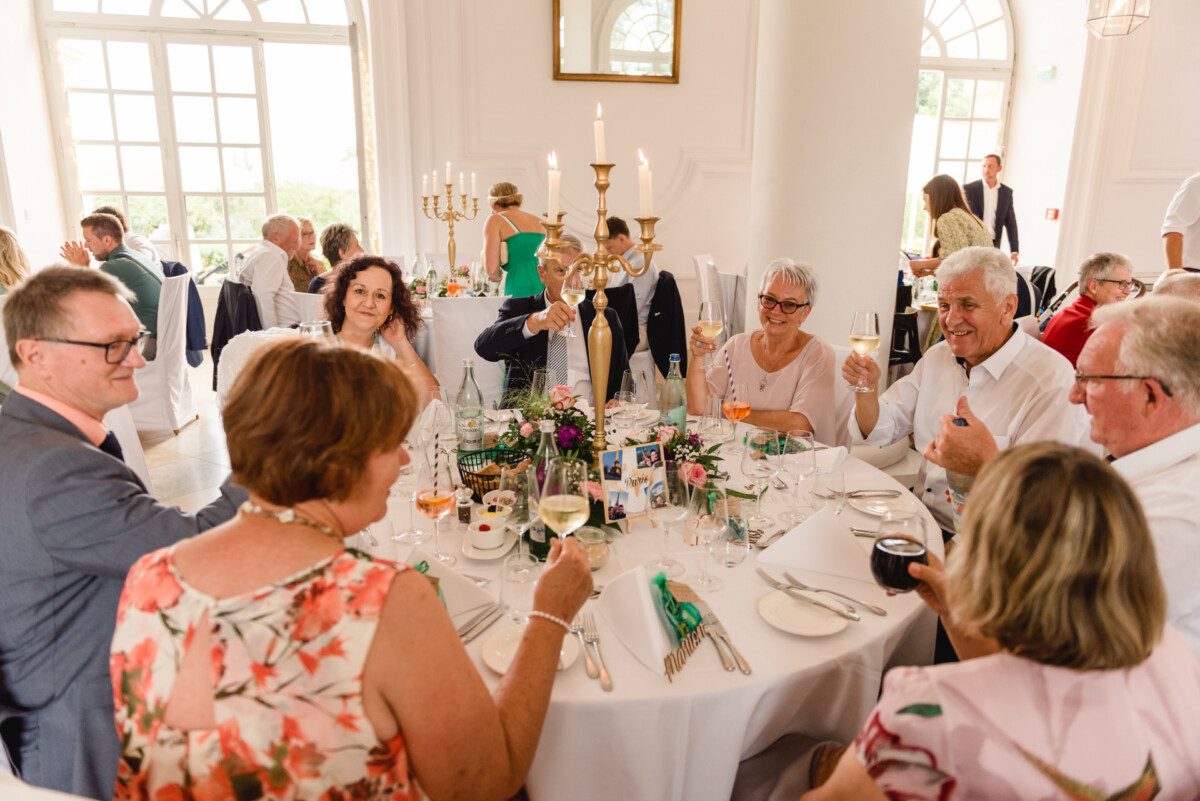 christian-lorenz.net Menschen sitzen bei einer formellen Veranstaltung lächelnd um einen Tisch und stoßen an. Der Tisch ist mit Blumen, Kerzen und Fotos dekoriert.