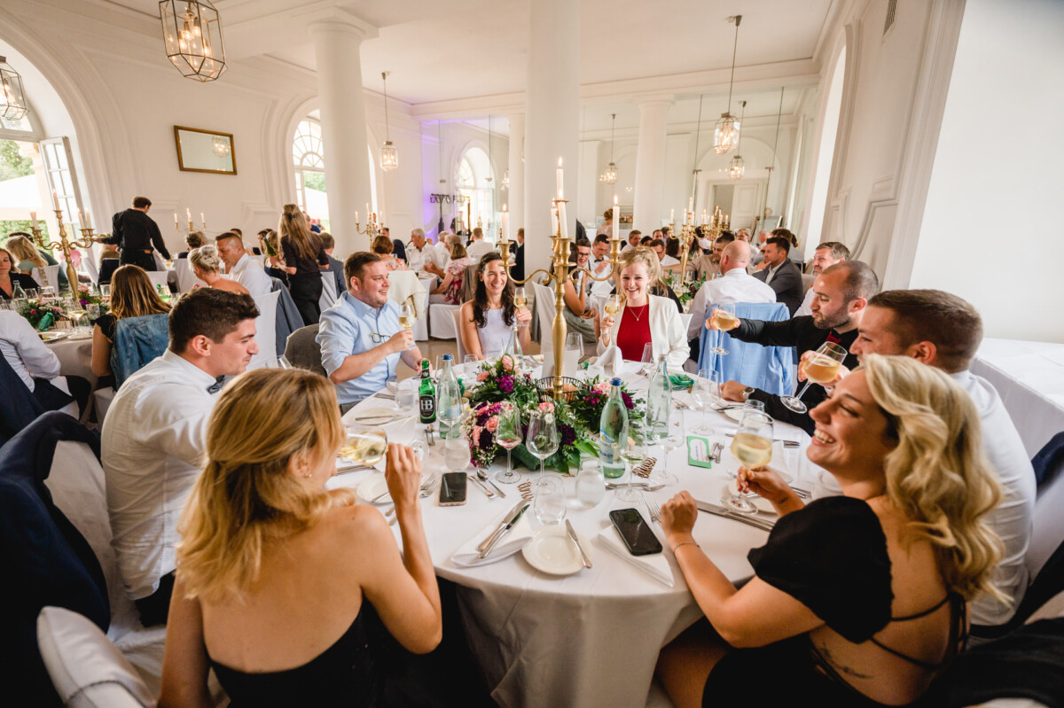 christian-lorenz.net Gäste sitzen an einem förmlich dekorierten Esstisch, halten Getränke hoch und lächeln, in einem eleganten Lokal mit weißen Säulen und großen Fenstern.