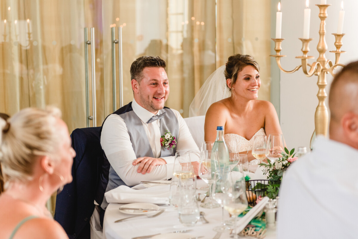 christian-lorenz.net Braut und Bräutigam sitzen während einer Hochzeitsfeier an einem Tisch. Beide lächeln. Der Tisch ist mit Gläsern, Tellern und einem Kerzenleuchter gedeckt.