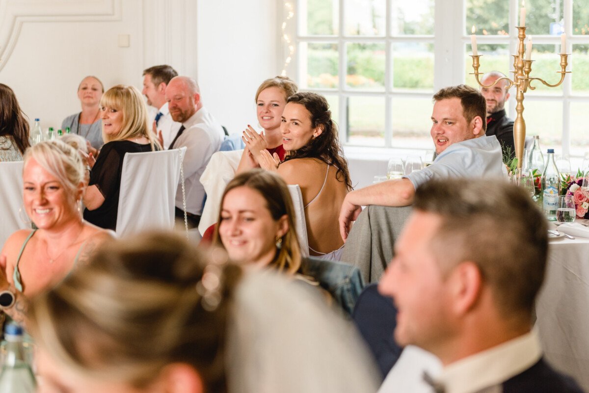 christian-lorenz.net Gäste sitzen bei einer formellen Veranstaltung an Tischen, lächeln und klatschen. Im Hintergrund sind ein großes Fenster und ein Kandelaber zu sehen.