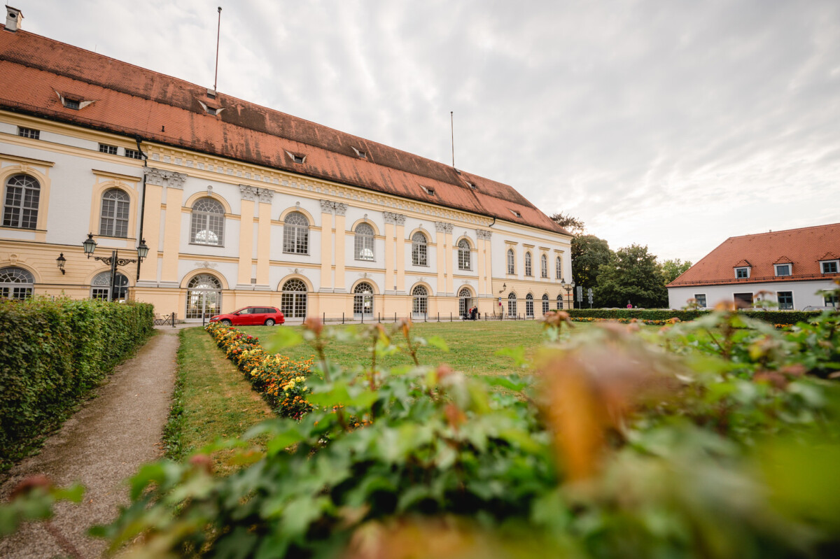 christian-lorenz.net Historisches Gebäude mit rotem Ziegeldach und hellgelber Fassade. Davor parkt ein rotes Auto. Im Vordergrund ist ein Garten mit Weg und grünen Hecken unter einem bewölkten Himmel zu sehen.