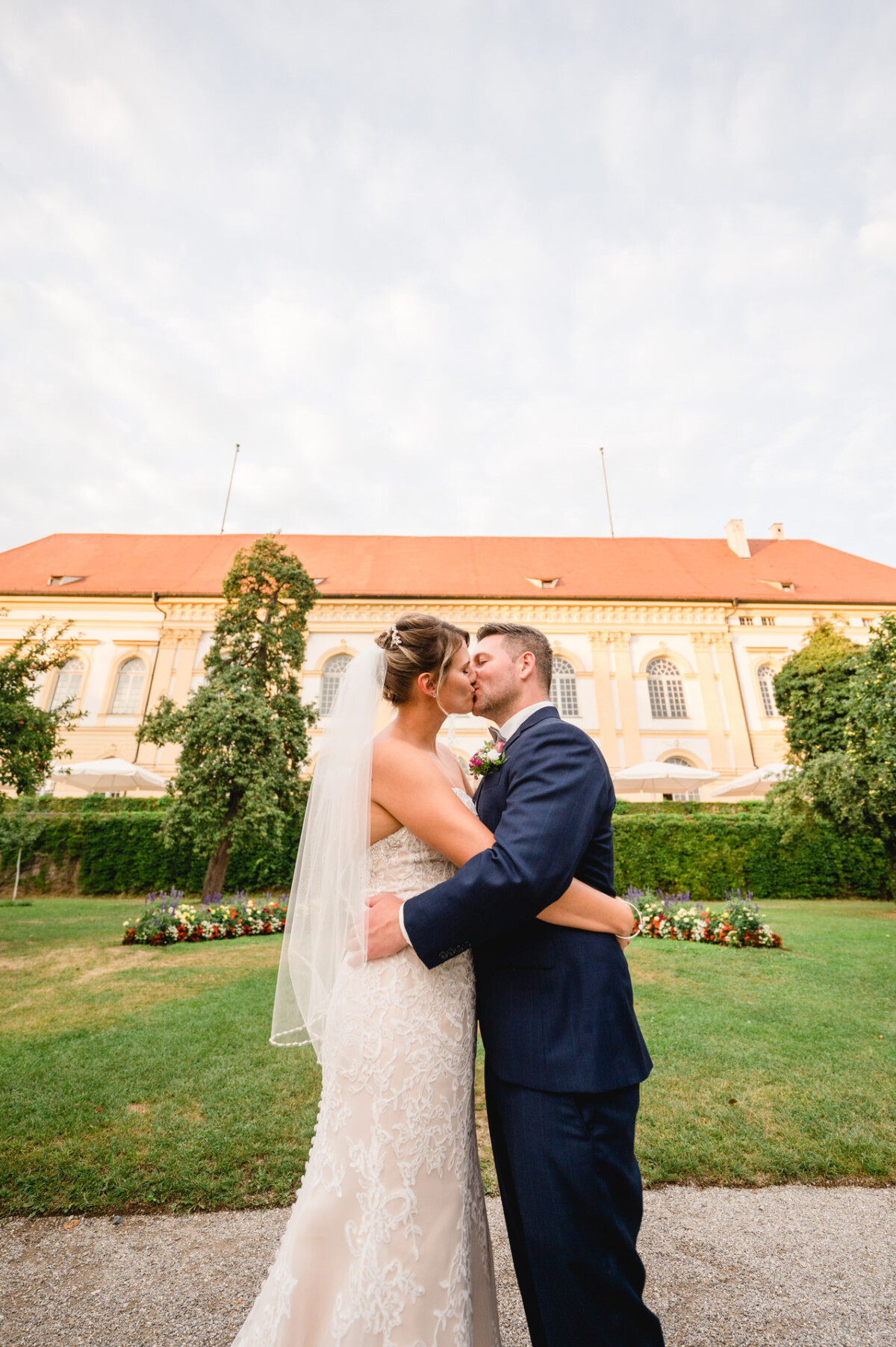 christian-lorenz.net Braut und Bräutigam umarmen und küssen sich vor einem großen, eleganten Gebäude mit roten Dächern. Die Kulisse sind gepflegte Gärten und ein klarer, teilweise bewölkter Himmel.