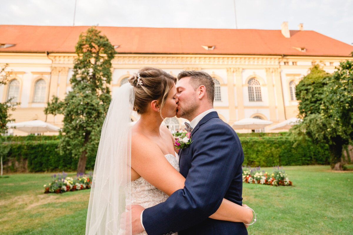 christian-lorenz.net Braut und Bräutigam küssen sich im Freien vor einem großen Gebäude mit rotem Dach und Bogenfenstern. Die Braut trägt einen Schleier, der Bräutigam einen dunklen Anzug.