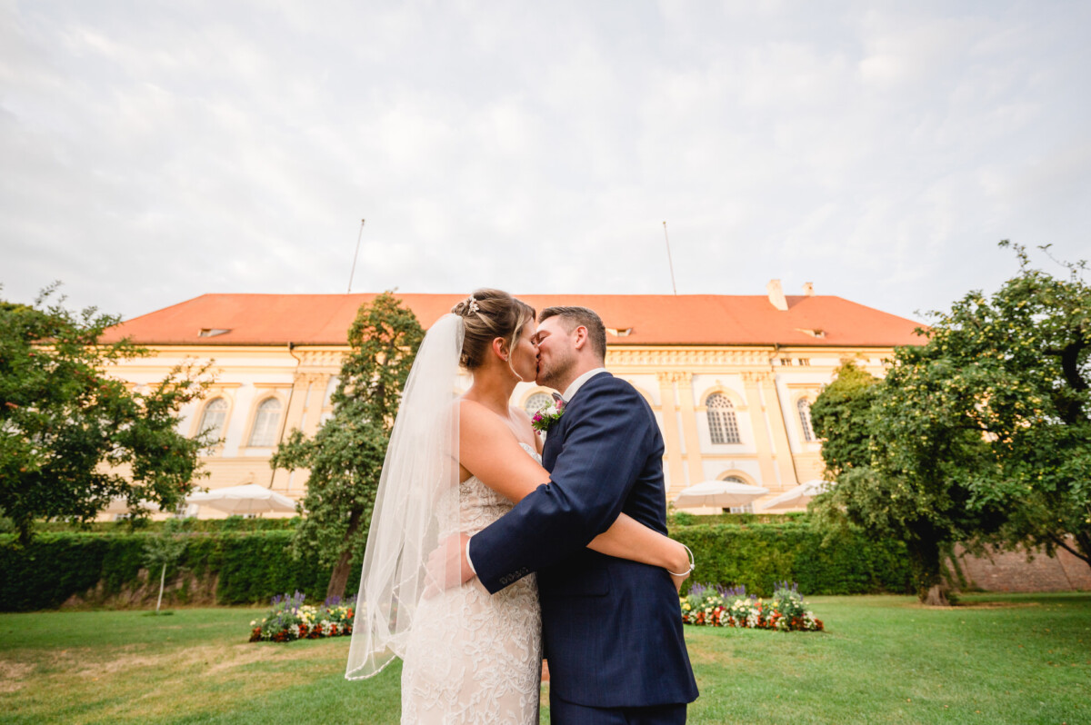 christian-lorenz.net Eine Braut und ein Bräutigam umarmen und küssen sich in einem Garten, im Hintergrund ist ein großes Gebäude mit einem roten Dach zu sehen.