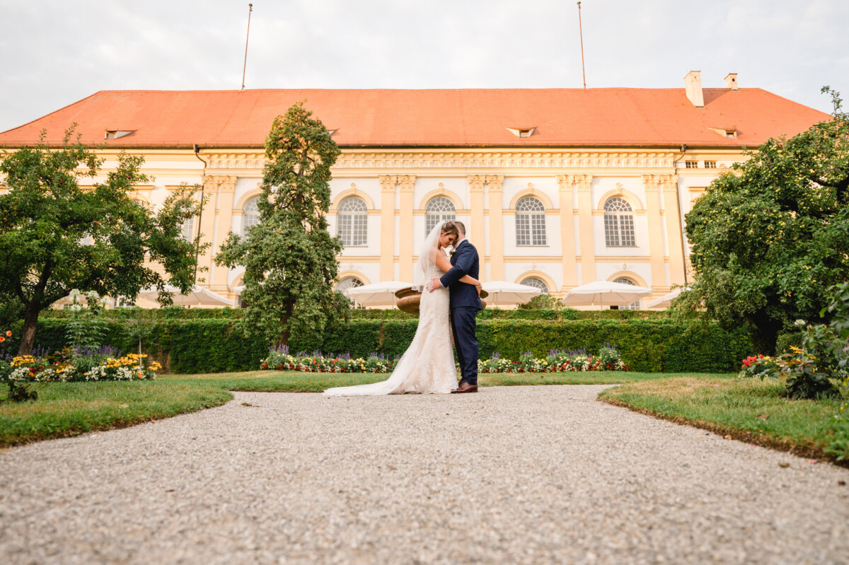 christian-lorenz.net Braut und Bräutigam umarmen sich auf einem Kiesweg vor einem großen, historischen Gebäude mit rotem Dach, umgeben von Grün und Blumen.