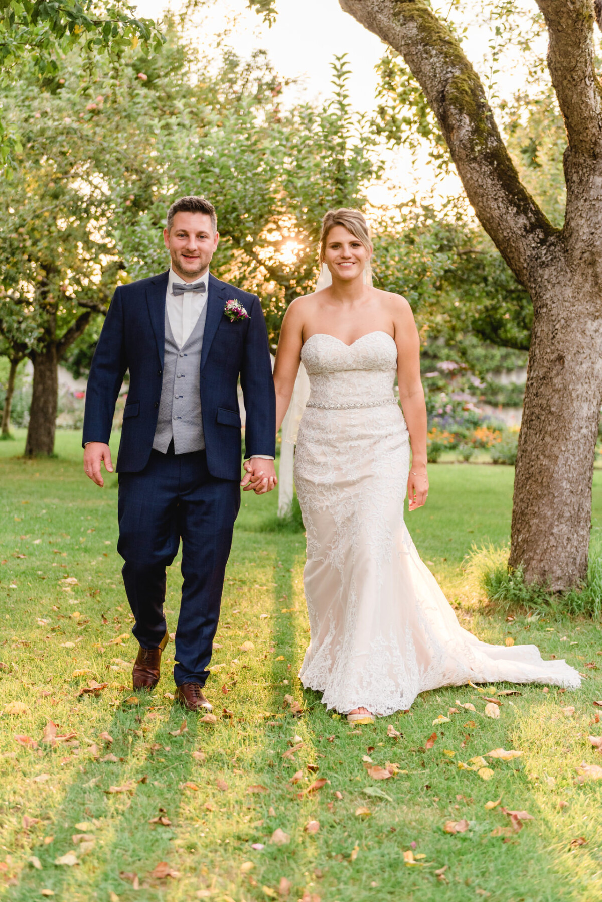 christian-lorenz.net Braut im weißen Kleid und Bräutigam im marineblauen Anzug gehen Hand in Hand auf einer Wiese, umgeben von Bäumen, durch die das Sonnenlicht fällt.