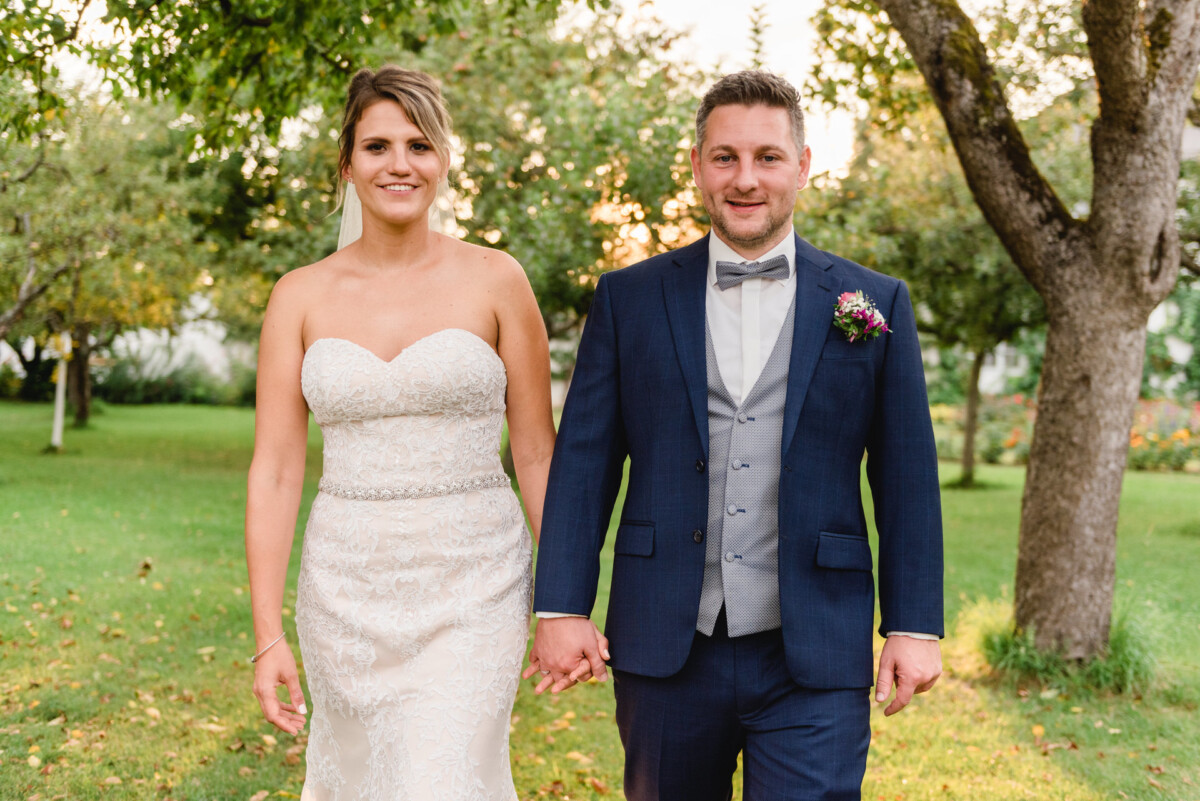 christian-lorenz.net Braut und Bräutigam stehen Händchen haltend im Freien. Die Braut trägt ein trägerloses weißes Kleid, der Bräutigam einen dunkelblauen Anzug mit Fliege. Sie lächeln in einer grünen Parklandschaft.